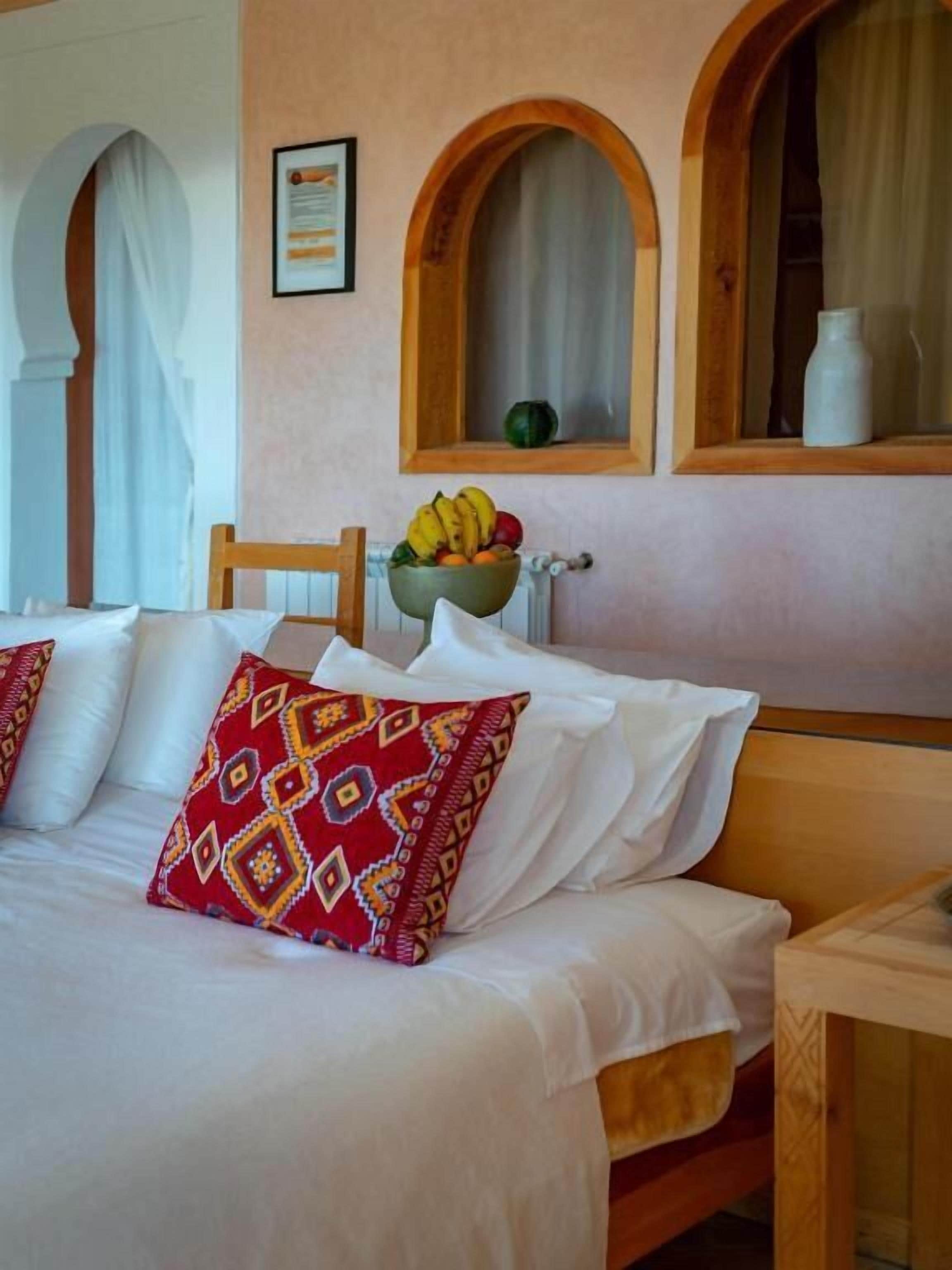 A hotel room with a bed, a wooden nightstand, a bowl of fruit, and a window with an arched frame.