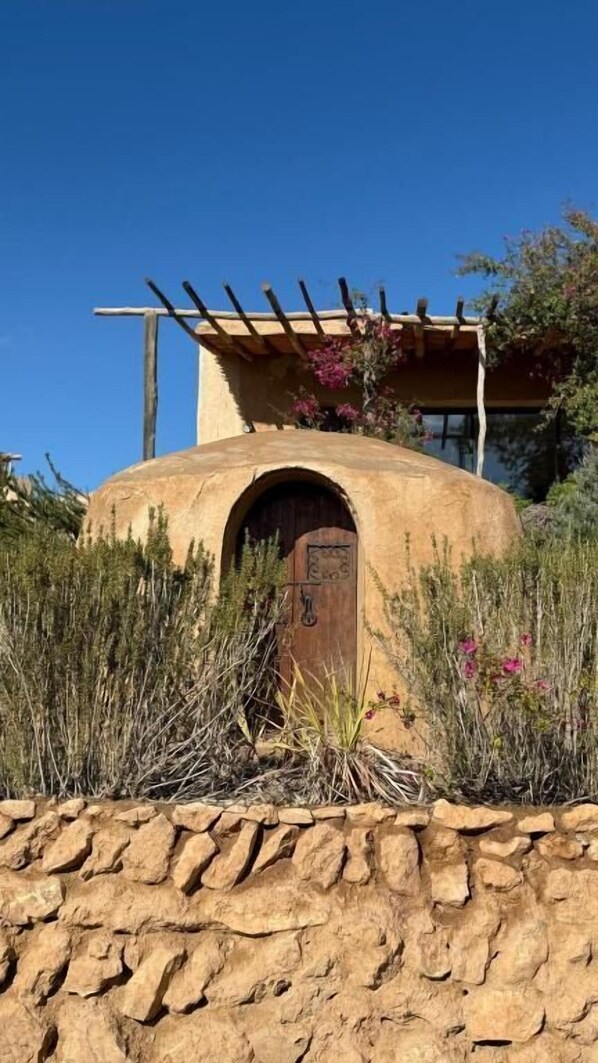 Edificio circular con puerta de madera, rodeado de vegetación y un muro de piedra.