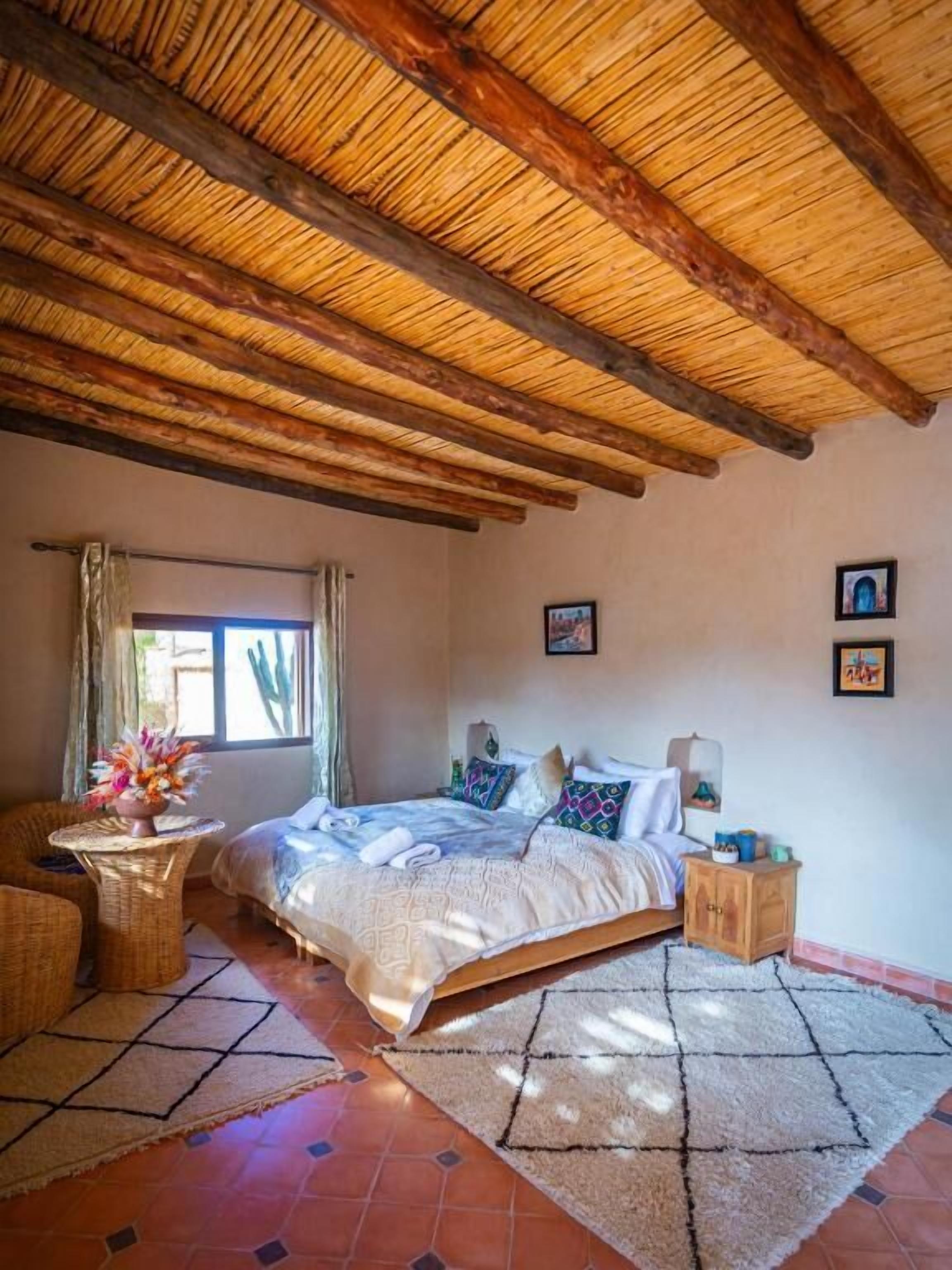 Une chambre à coucher avec un lit, une table de chevet, un fauteuil en osier et une fenêtre avec des rideaux.