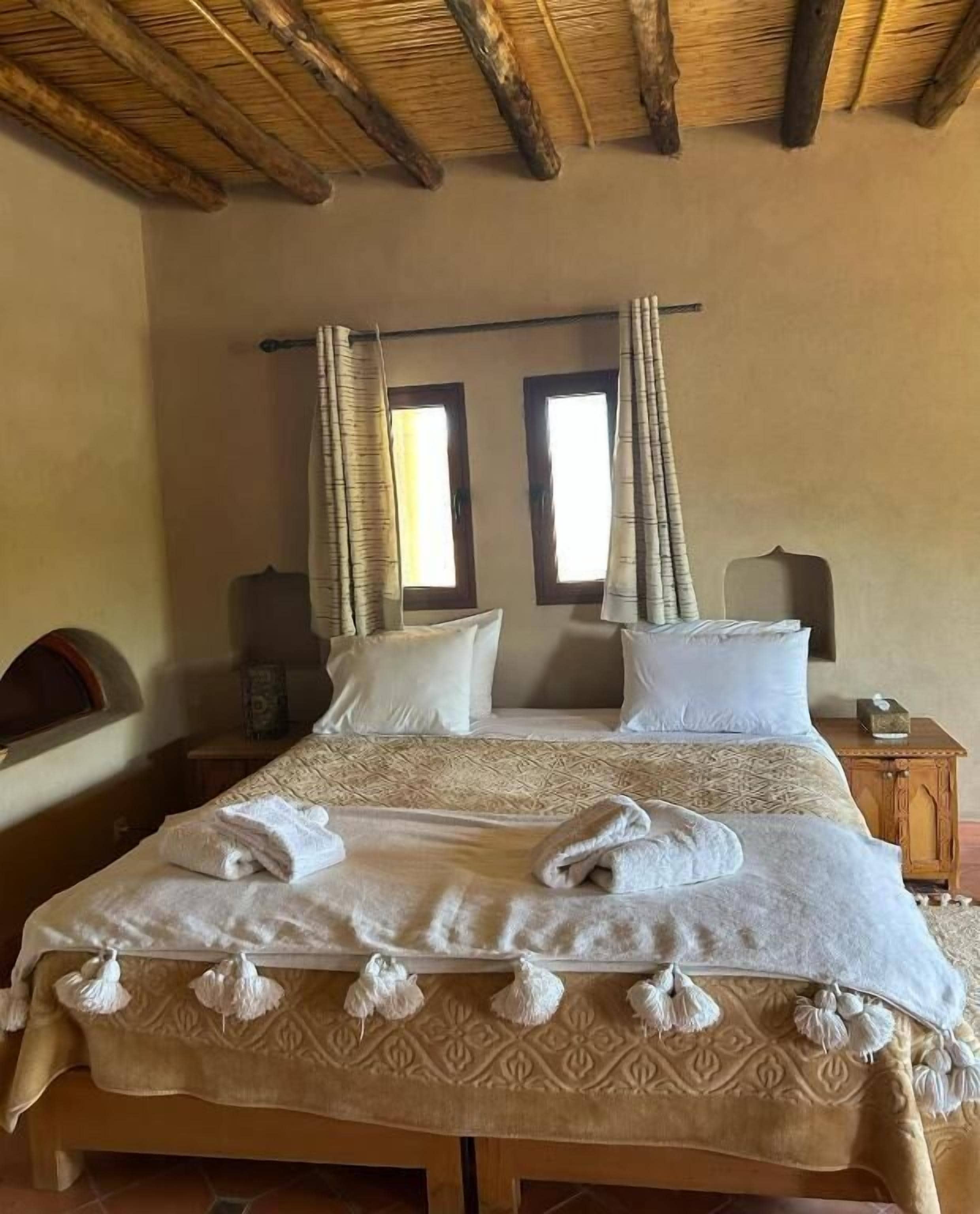 Un letto matrimoniale con una coperta a motivi decorativi e due cuscini, un comodino con una lampada e una finestra con tende.