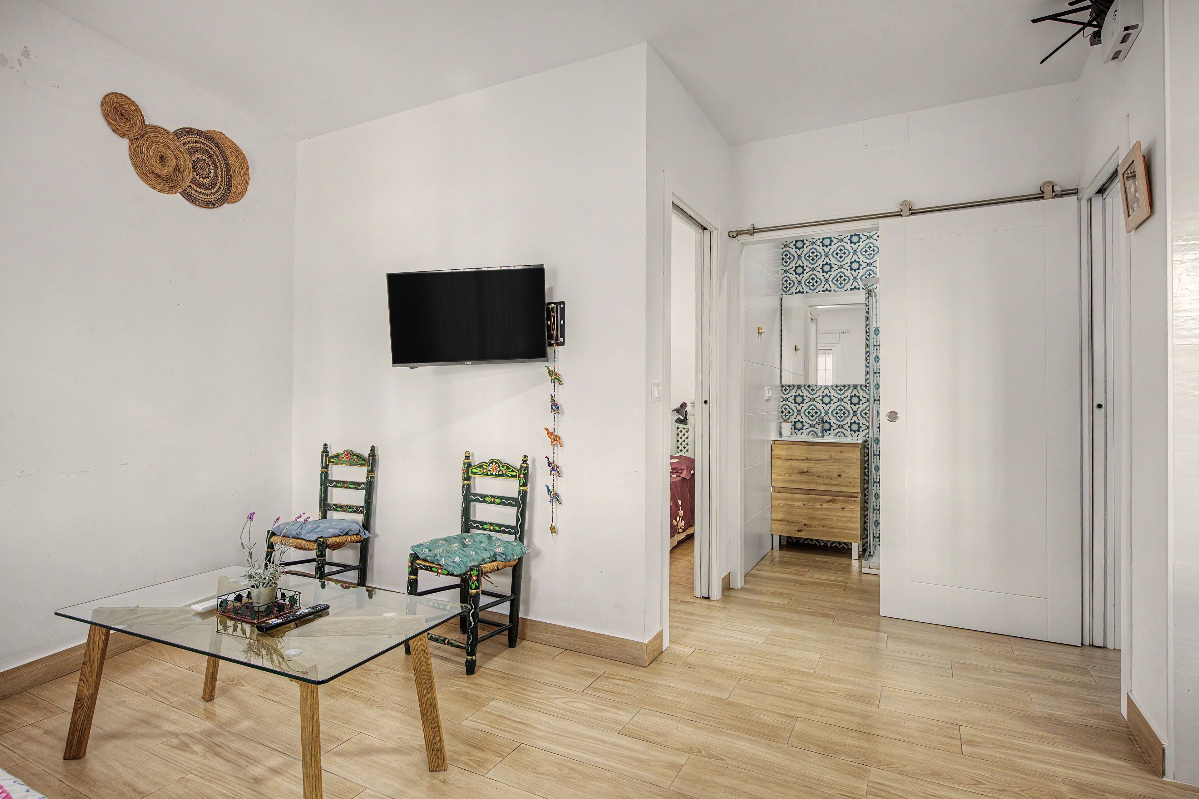 A room with a television mounted on the wall, two chairs with cushions, a glass-top table with a remote control, and a wooden floor.