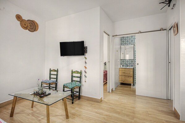 Ein Zimmer mit einem an der Wand befestigten Fernseher, zwei Sessel mit Kissen, einem Glastisch und einer Fernbedienung sowie einem Holzboden.