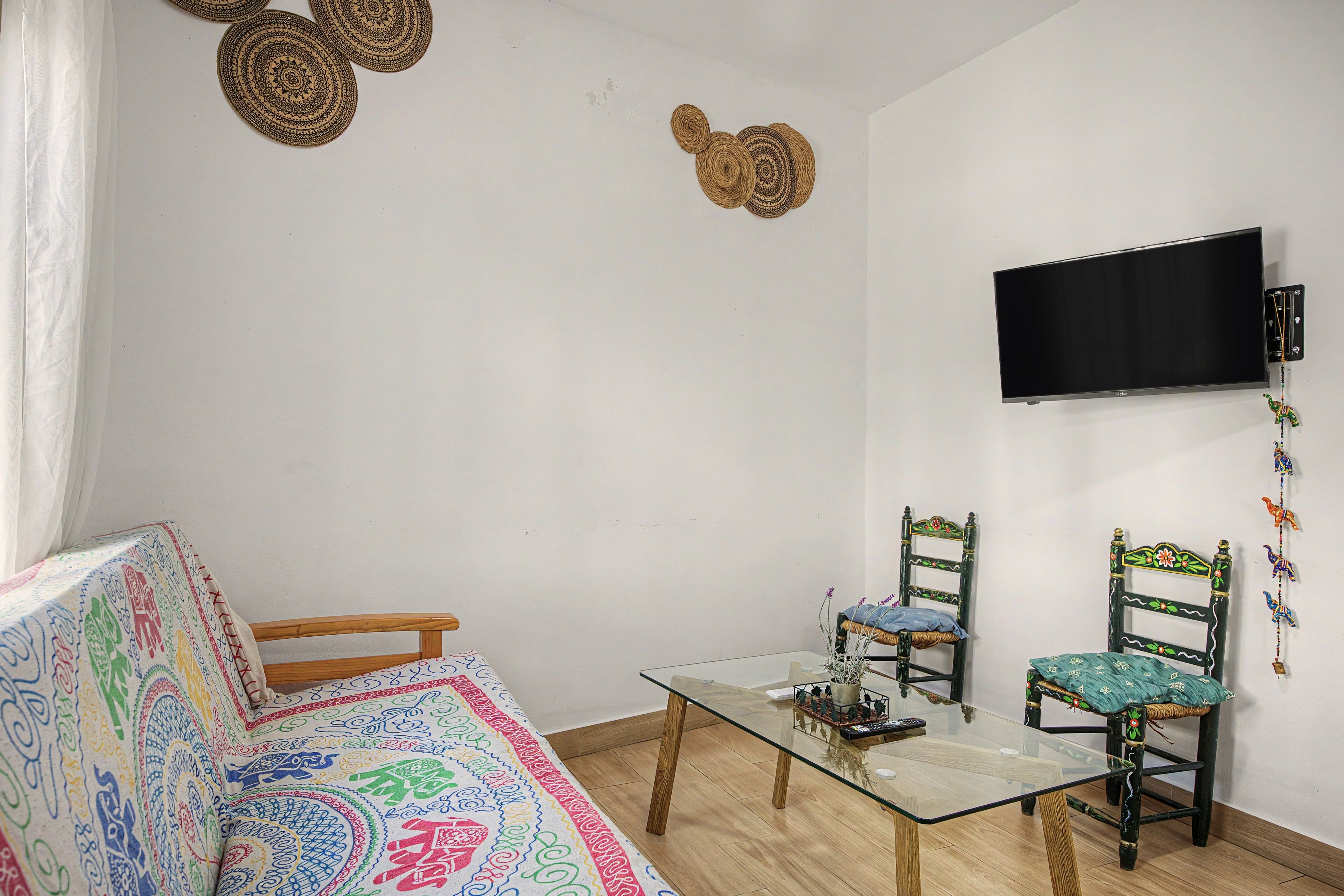 A room with a glass-top table, two chairs, a sofa, a television mounted on the wall, and decorative wall hangings.