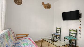 A room with a glass-top table, two chairs, a sofa, a television mounted on the wall, and decorative wall hangings.