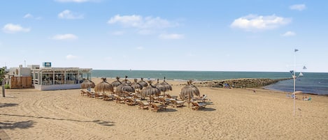 A beach with sun loungers and umbrellas, a building with a sign, and a rocky shoreline.