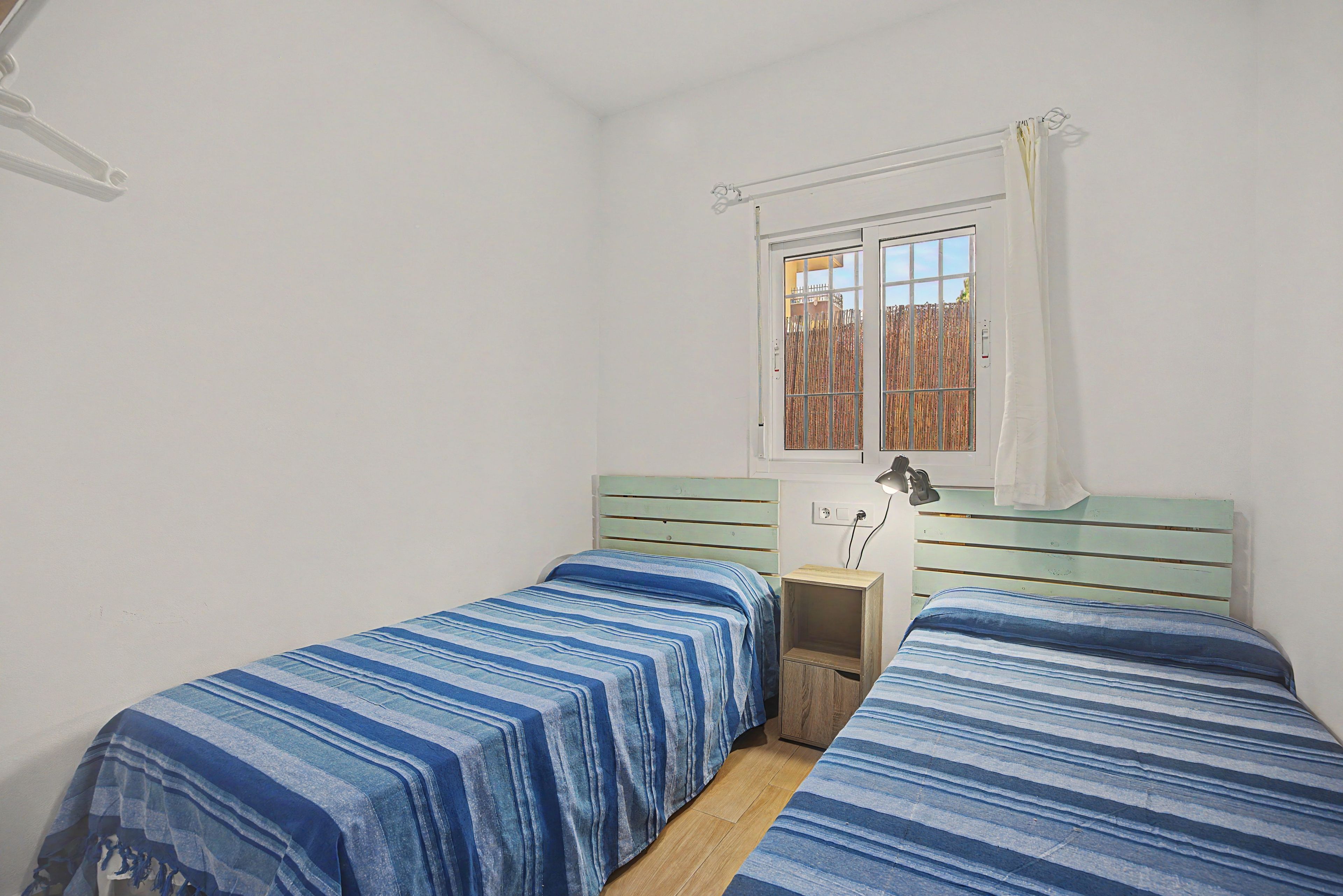 Ein Schlafzimmer mit zwei Einzelbetten, einem Fenster mit Blick auf einen Zaun und einem kleinen Nachttisch mit einer Lampe.