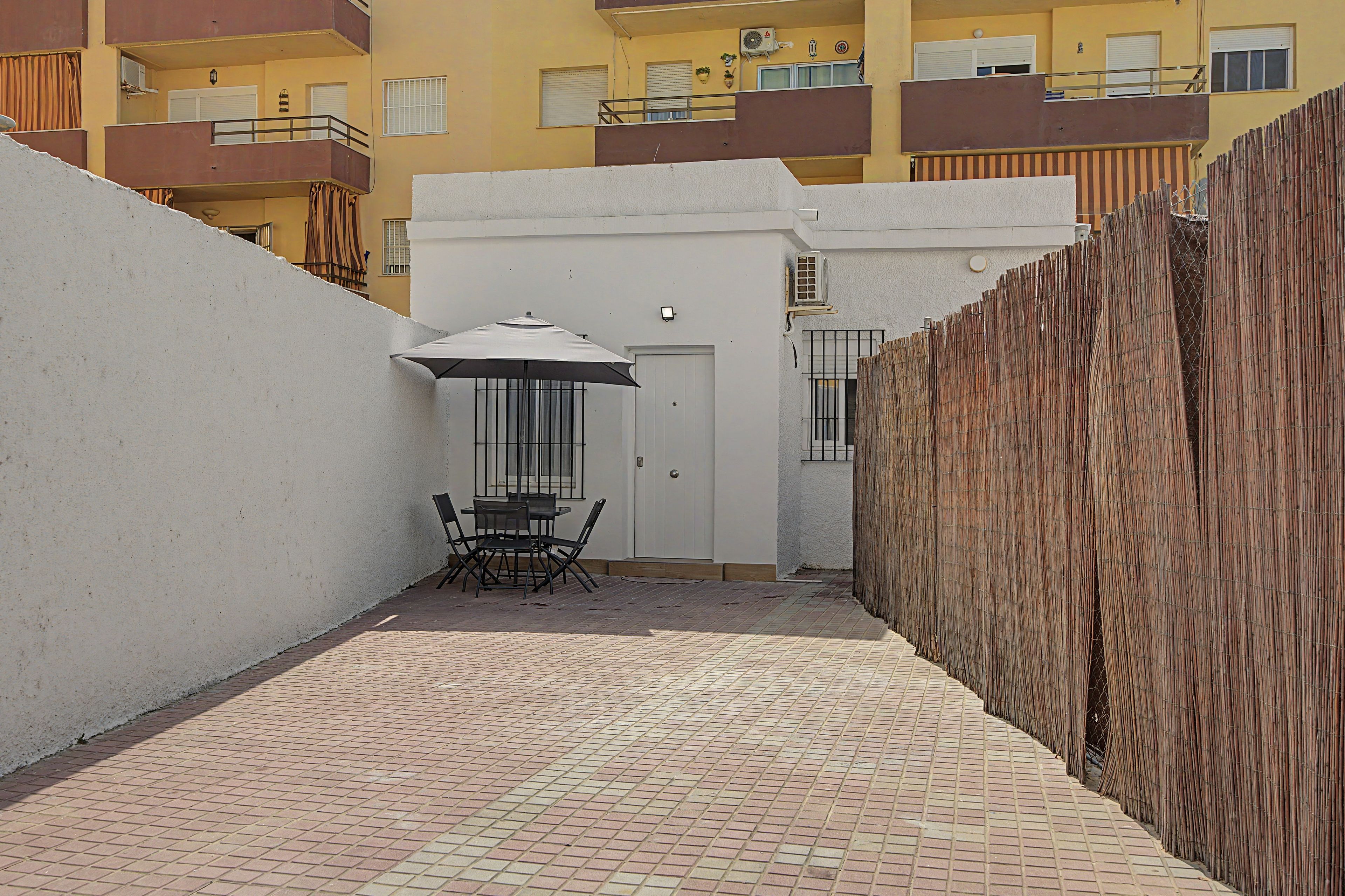 A narrow courtyard with a table and chairs, a door, and a small covered area with an umbrella.