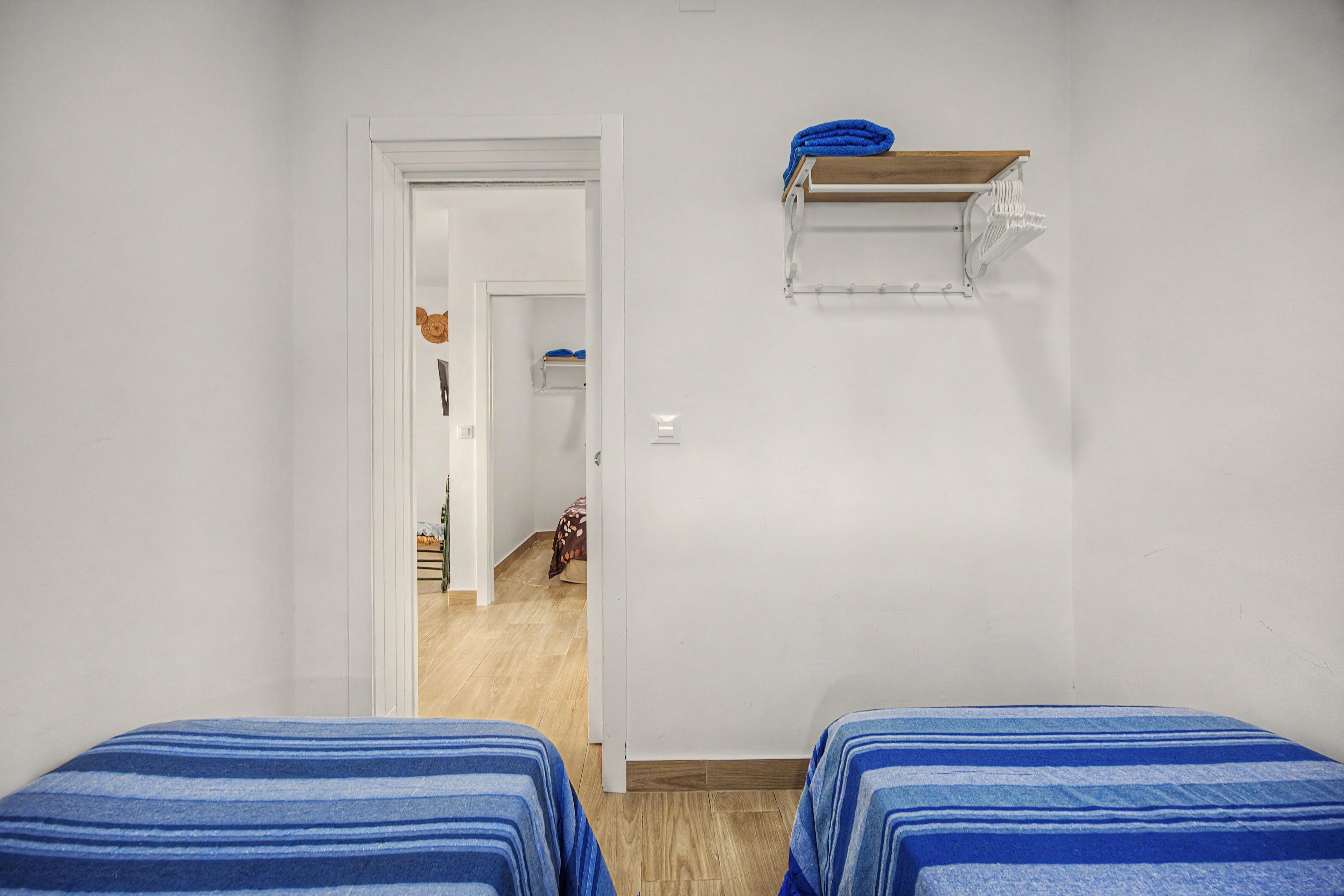 A room with two beds, a wooden floor, a white door, and a small shelf with a towel.