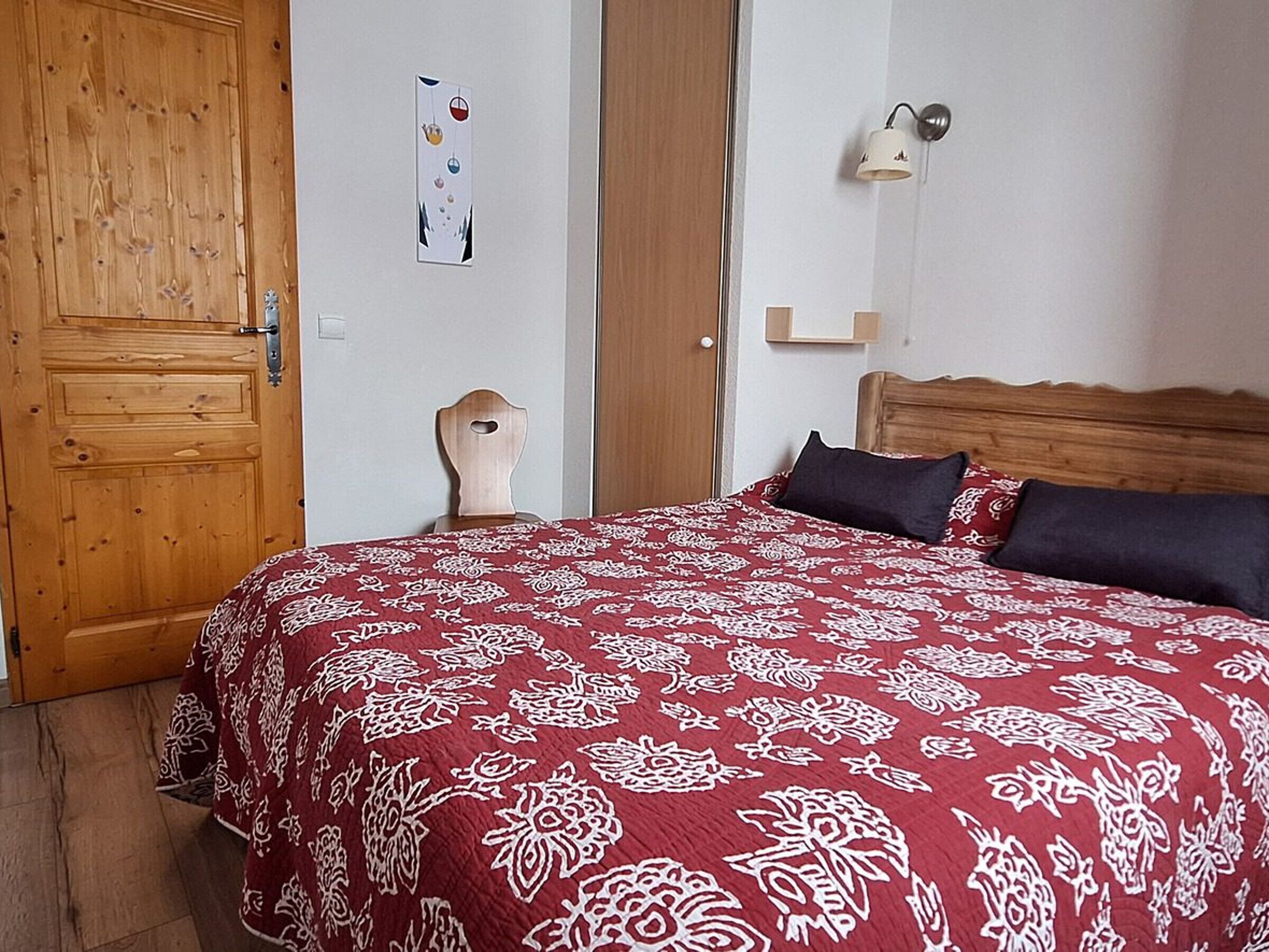 A bedroom with a wooden door, a bed with a patterned bedspread, a bedside lamp, and a wall-mounted picture.