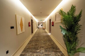 A hotel corridor with a carpeted floor, wall-mounted fire extinguishers, and a potted plant on the right.