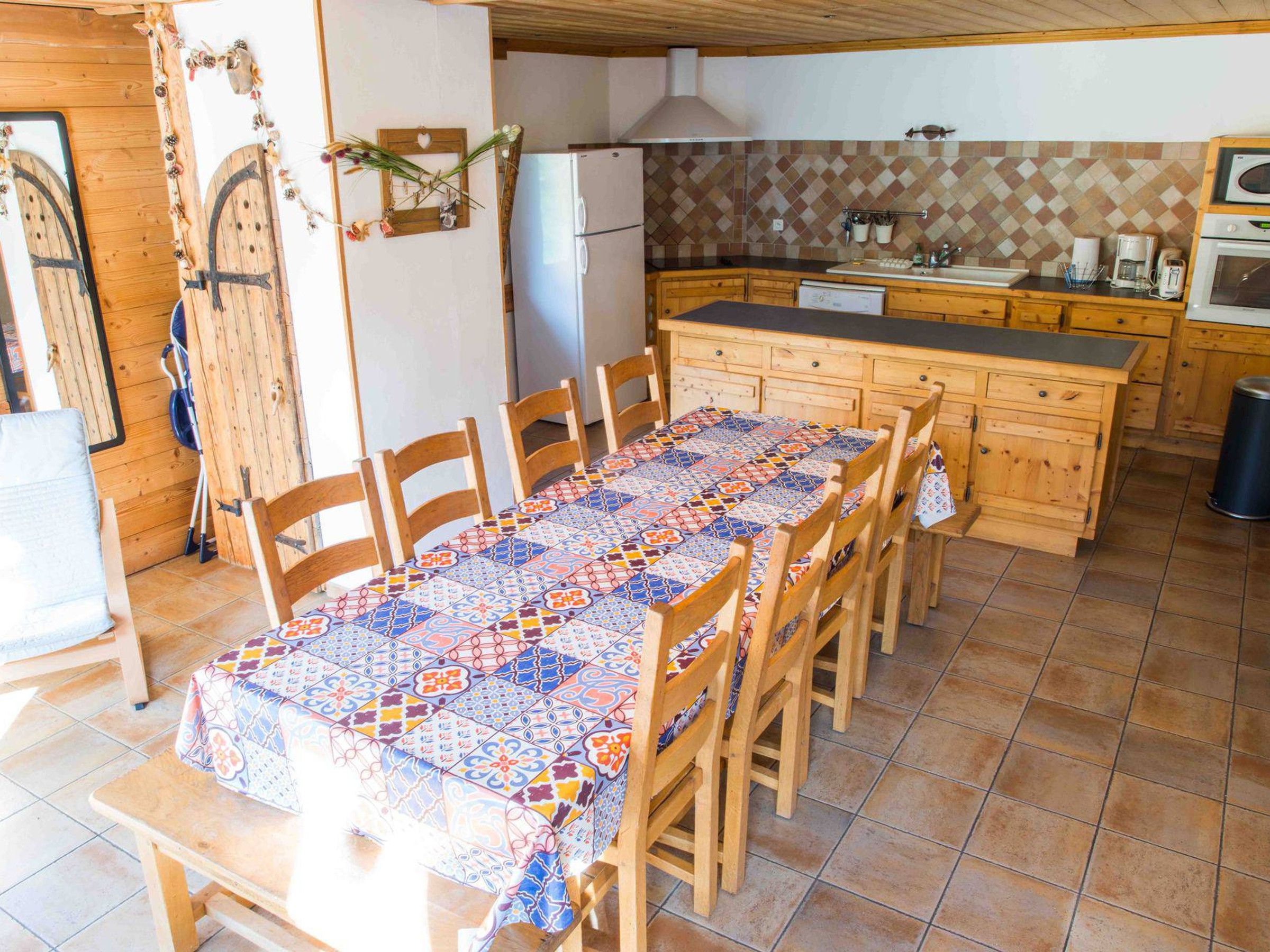 Una cocina con salpicadero de azulejos, armarios de madera, un refrigerador y un comedor con mantel estampado.