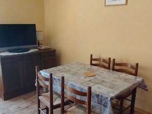 Ein Essbereich mit einem Tisch, der mit einer gemusterten Tischdecke bedeckt ist, vier Holzstühlen, einem Fernseher auf einem Schrank und einer Lampe auf dem Tisch.