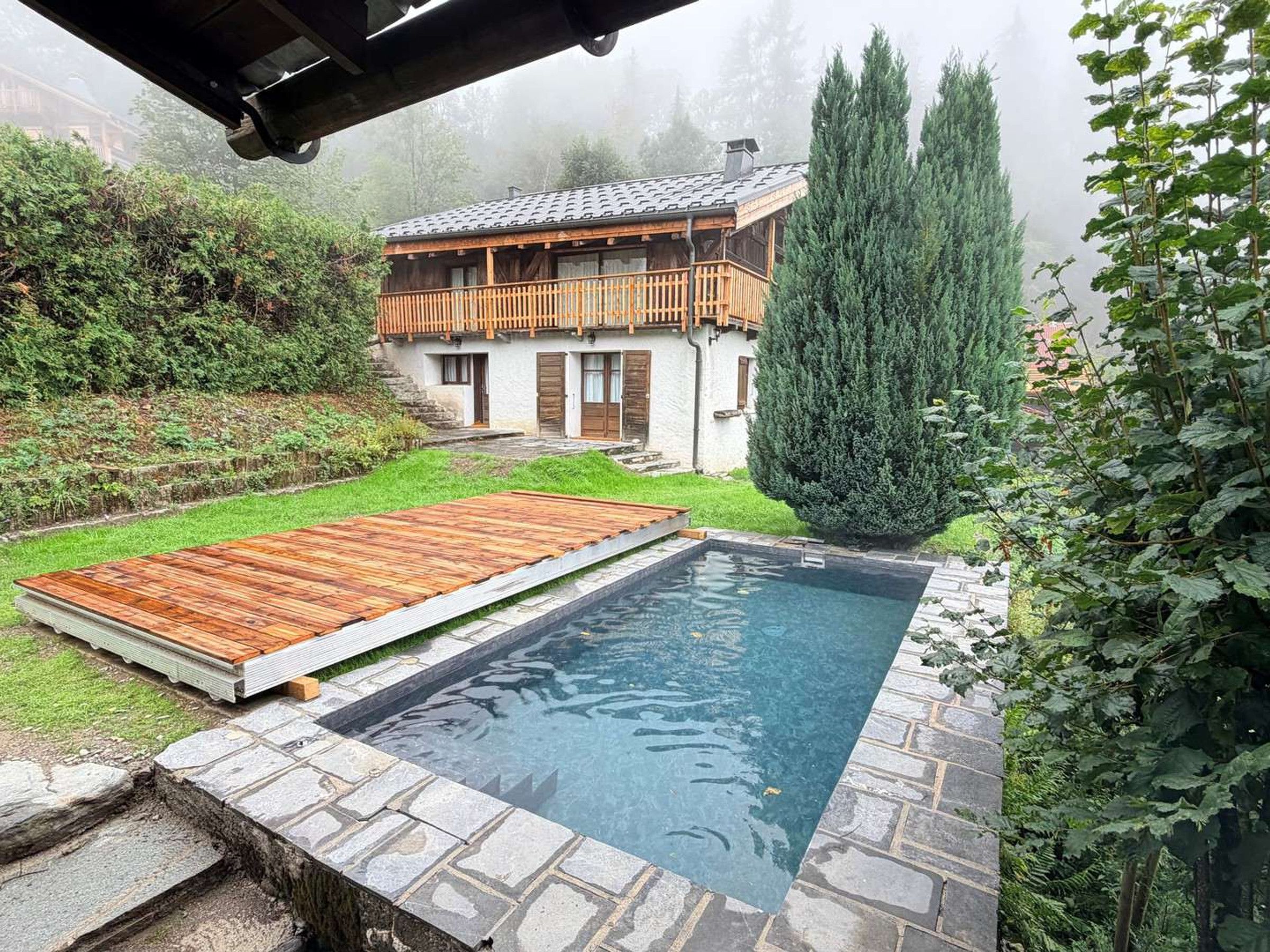 A swimming pool with a wooden deck, surrounded by a stone border, and a house with a balcony in the background.