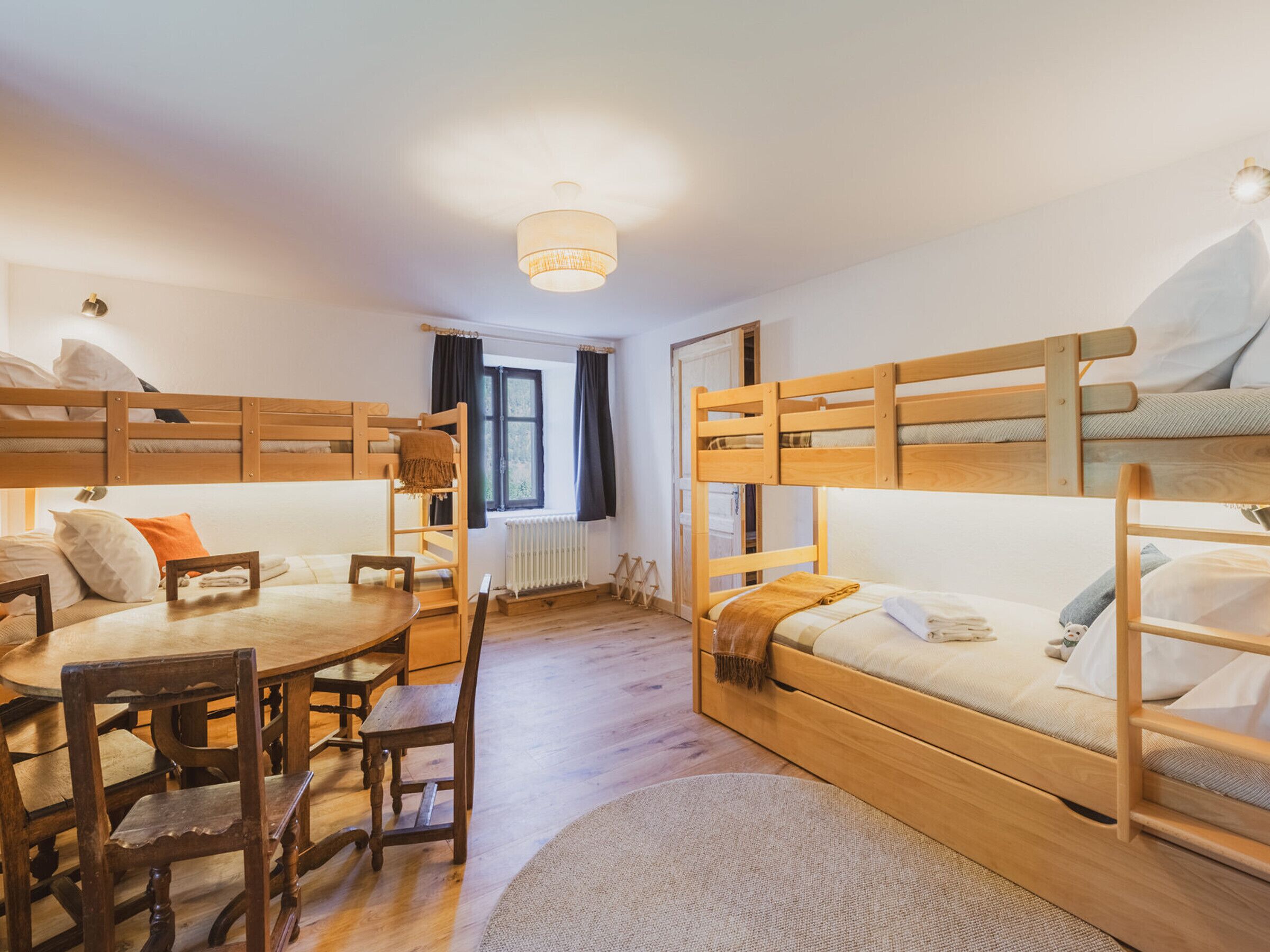 Une chambre de dortoir avec des lits superposés, une table en bois, des chaises, un radiateur et une fenêtre avec des rideaux.
