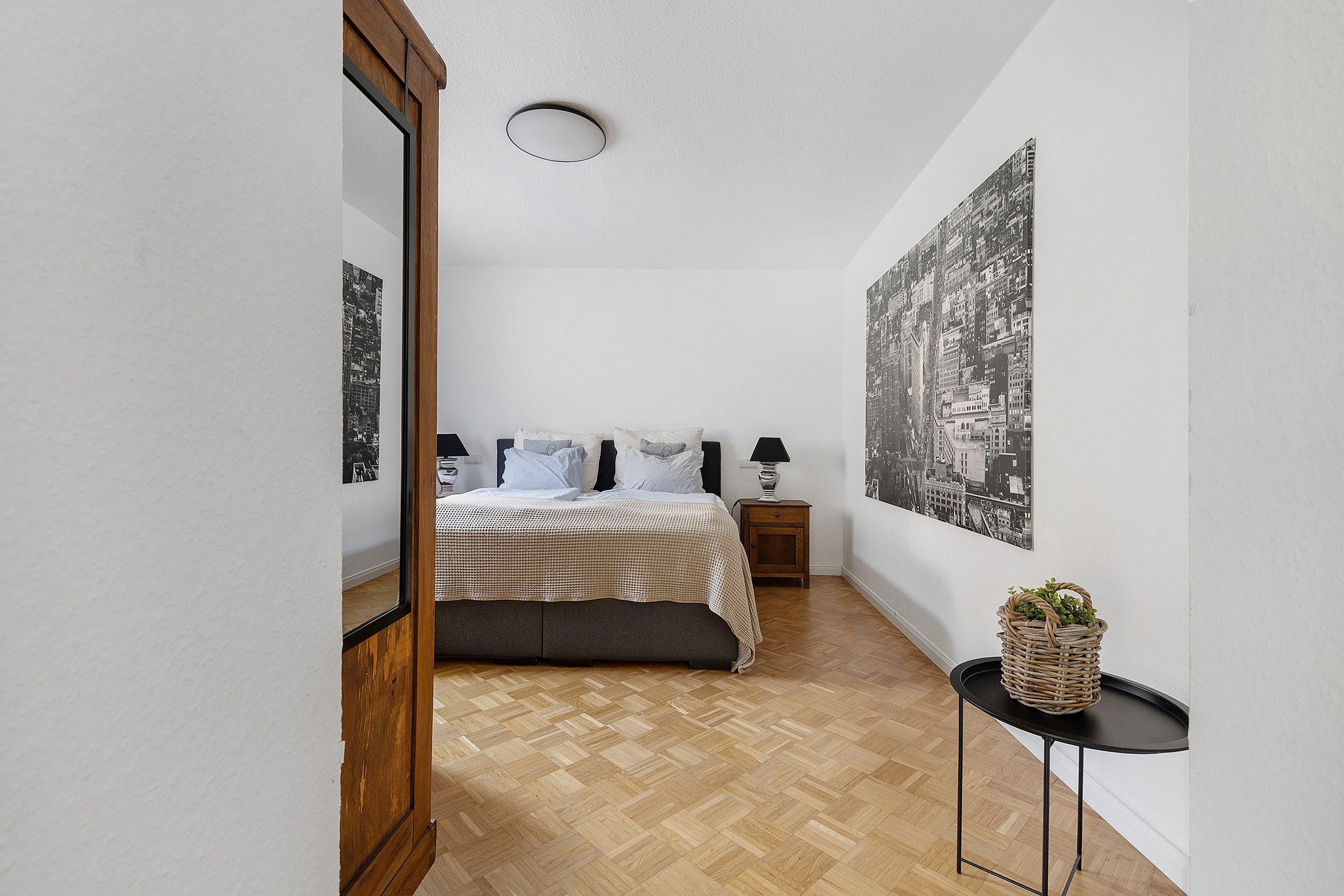 A bedroom with a bed, a nightstand, a lamp, a wall-mounted picture, and a small table with a plant.