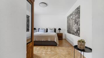 A bedroom with a bed, a nightstand, a lamp, a wall-mounted picture, and a small table with a plant.