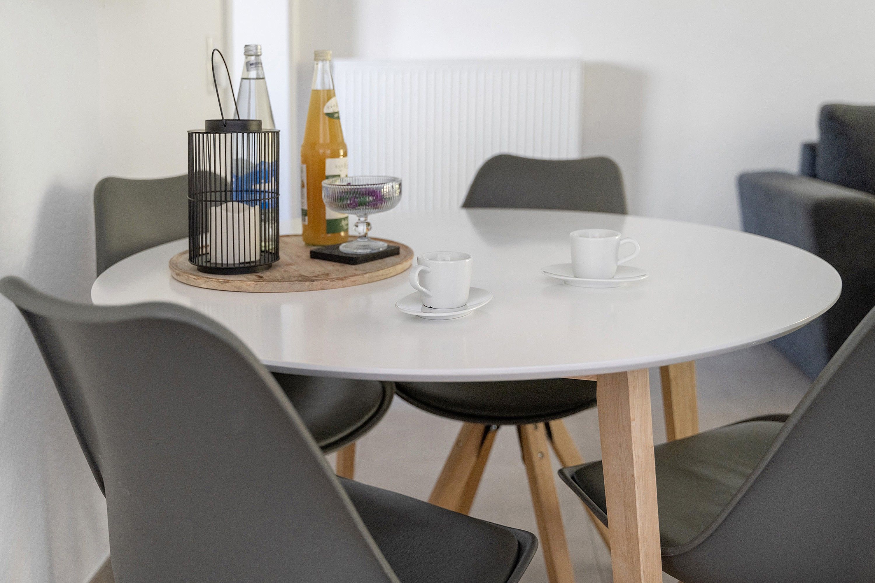 Ein runder Tisch mit weißer Tischplatte, umgeben von vier grauen Stühlen mit Holzbeinen. Auf dem Tisch stehen zwei Flaschen, eine Glaskugel mit violettem Inhalt und zwei weiße Tassen auf Untertellern.