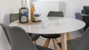 Ein runder Tisch mit weißer Tischplatte, umgeben von vier grauen Stühlen mit Holzbeinen. Auf dem Tisch stehen zwei Flaschen, eine Glaskugel mit violettem Inhalt und zwei weiße Tassen auf Untertellern.