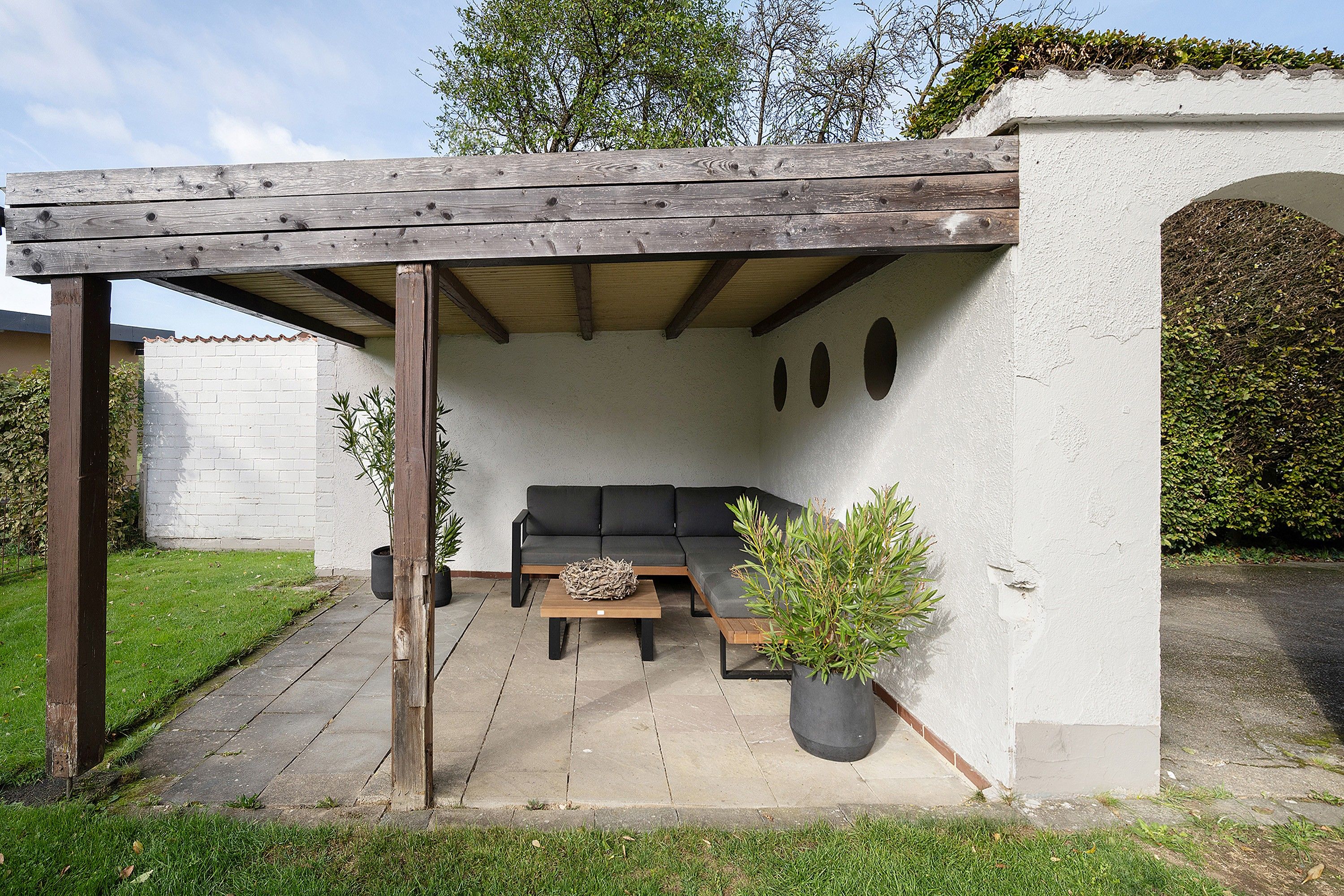 A covered outdoor seating area with a sofa and a coffee table, surrounded by greenery and a white wall.