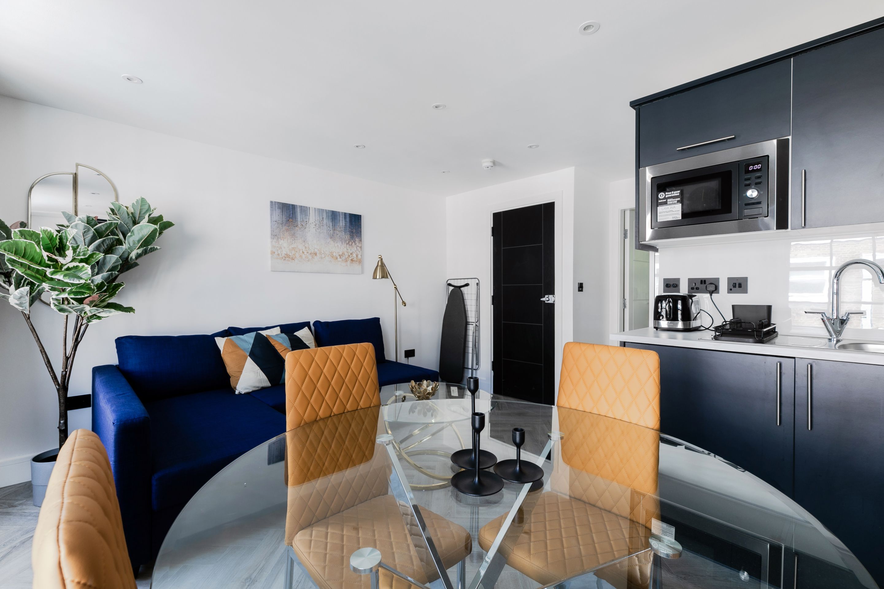 Una sala de estar moderna con un sofá azul, una mesa de comedor de vidrio y una zona de cocina con armarios oscuros y un microondas.