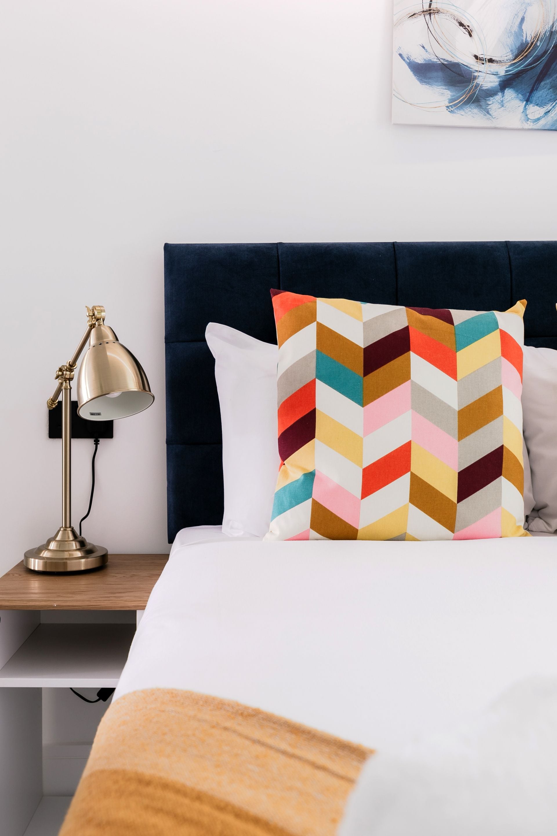 Una cama bien hecha con un cojín de estampado geométrico y llamativos colores, una lámpara de escritorio dorada y un cuadro abstracto colgado en la pared.