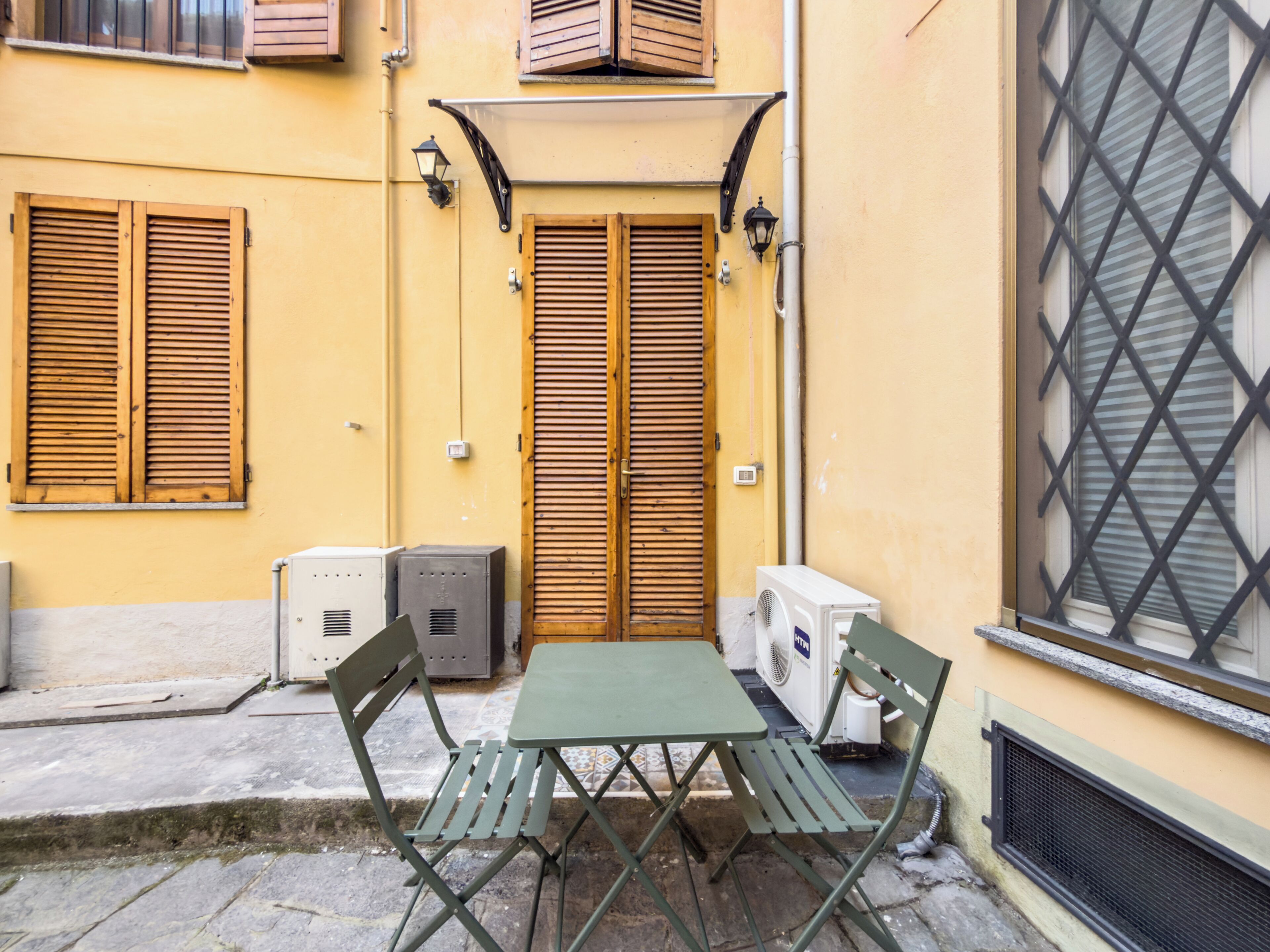 Ein kleiner Sitzbereich im Freien mit Tisch und Stühlen, ein Fenster mit Metallgitter und eine Tür mit Holzläden.