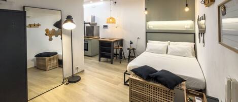 A modern hotel room with a bed, bedside table, lamp, and a view of the kitchen area.