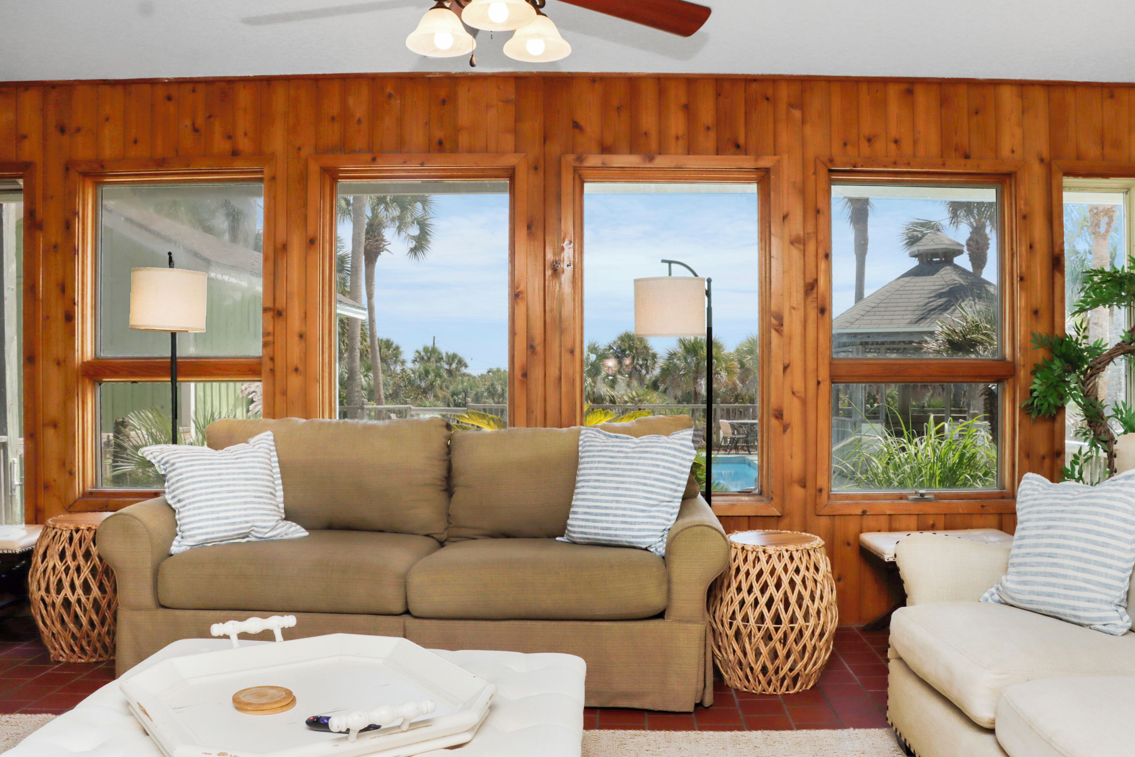 A room with a wooden paneled wall, a beige sofa with striped pillows, two side tables, and large windows with views of palm trees and a pool.