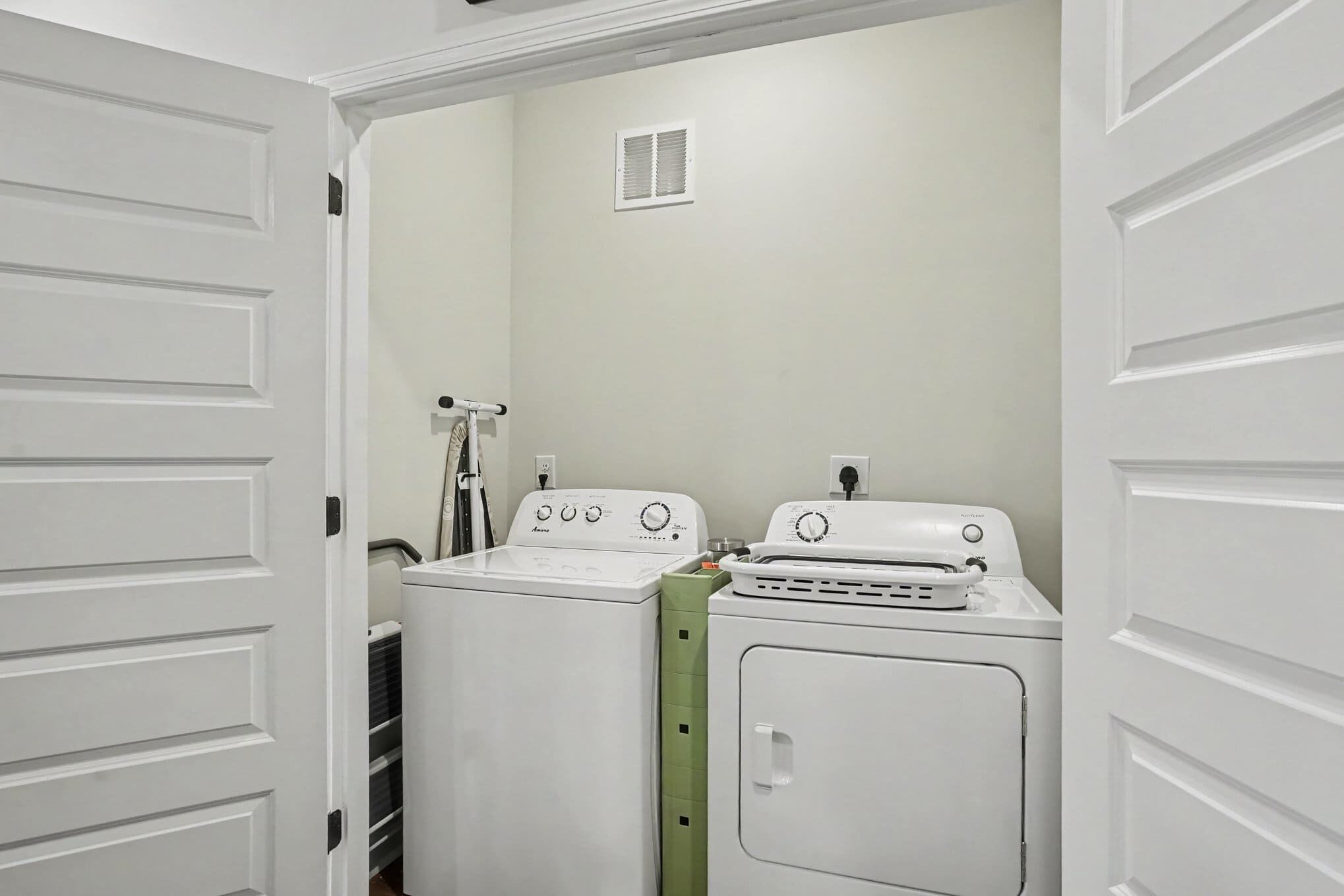 A laundry room with two washing machines and a dryer, a ventilation vent on the wall, and a door leading to another room.