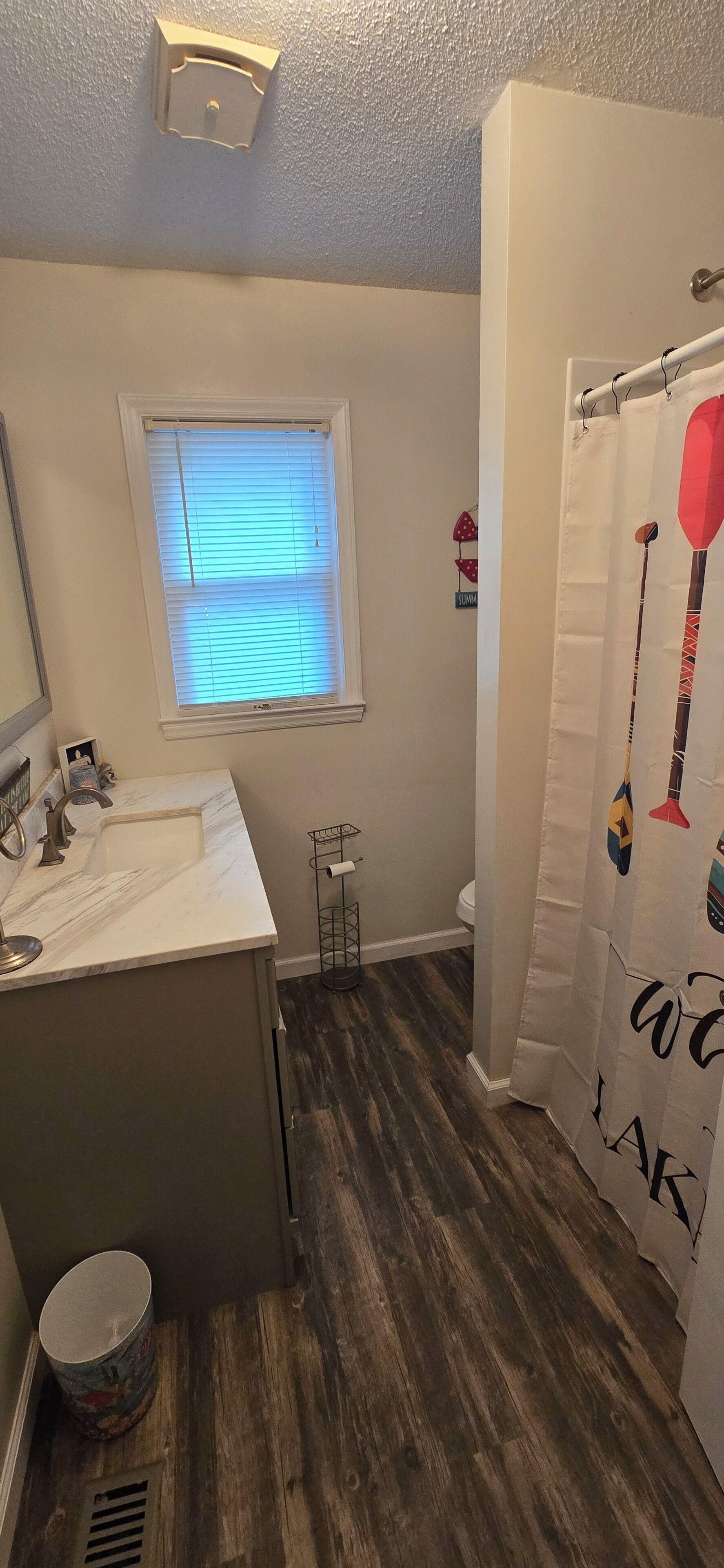 Un baño con un lavamanos blanco, una ventana con persianas y una cortina de ducha con diseños tribales, sobre un piso de madera.
