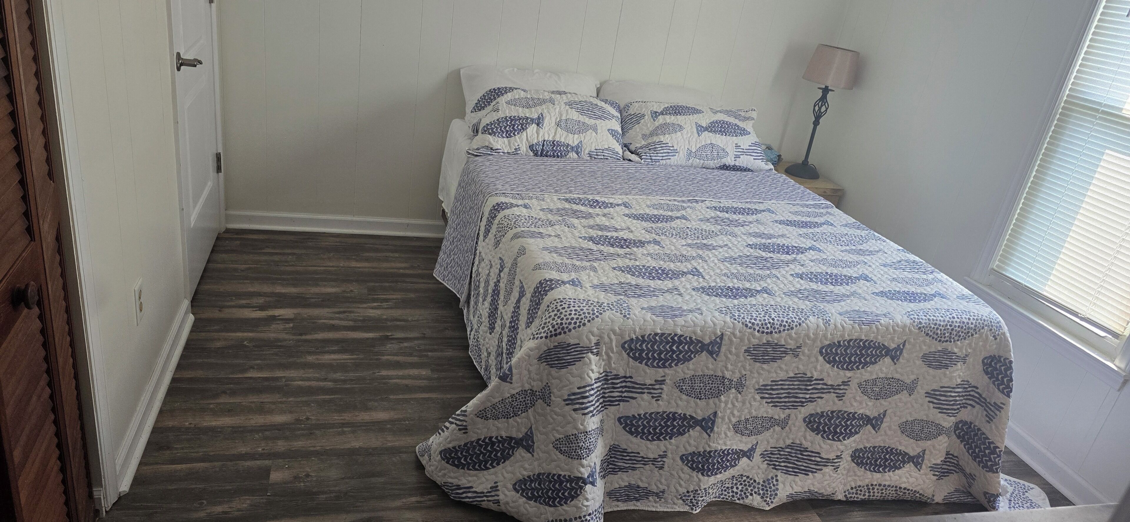 A single bed with a patterned bedspread, a bedside lamp, and a window with blinds.