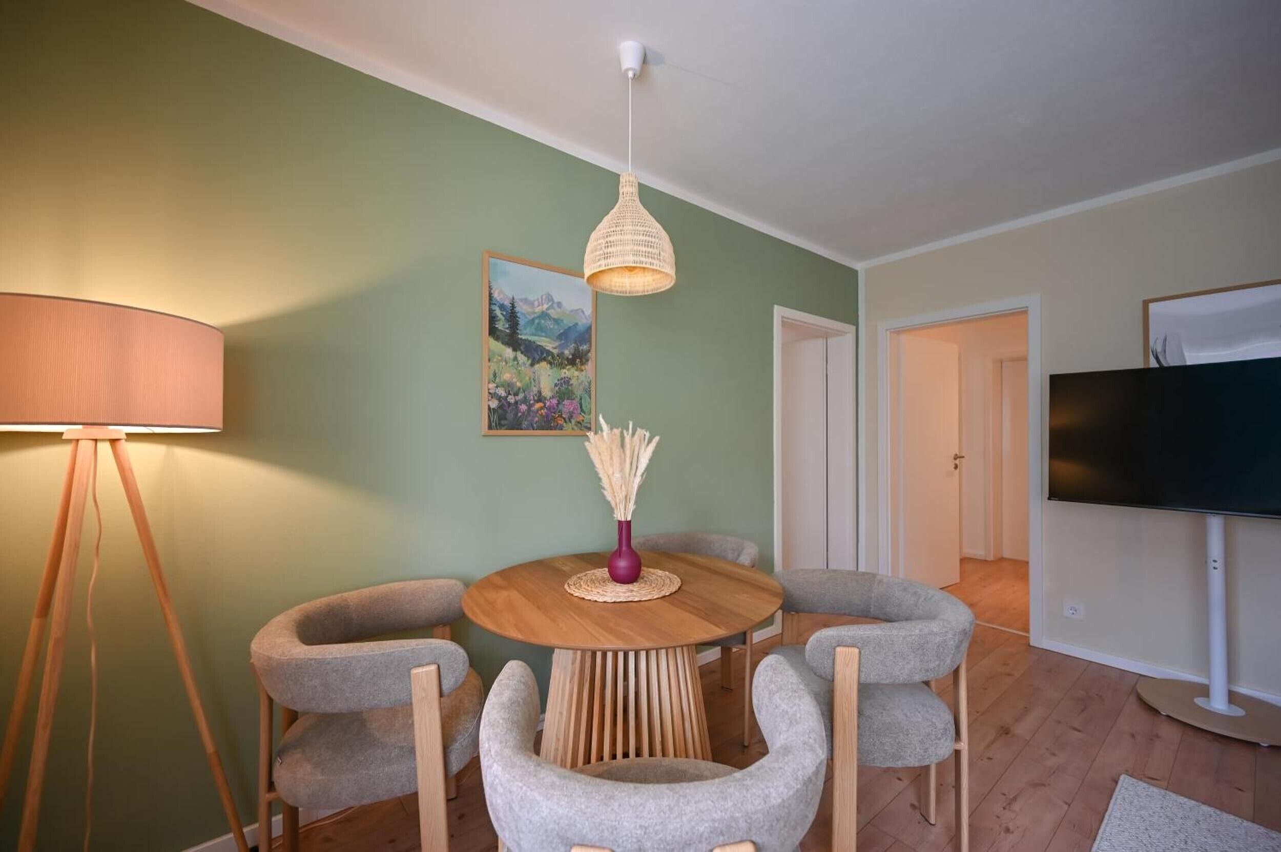 Ein Essbereich mit einem runden Tisch, vier grauen Stühlen, einer geflochtenen Pendelleuchte, einer Vase mit Trockenpflanzen und einem Bild an der Wand.