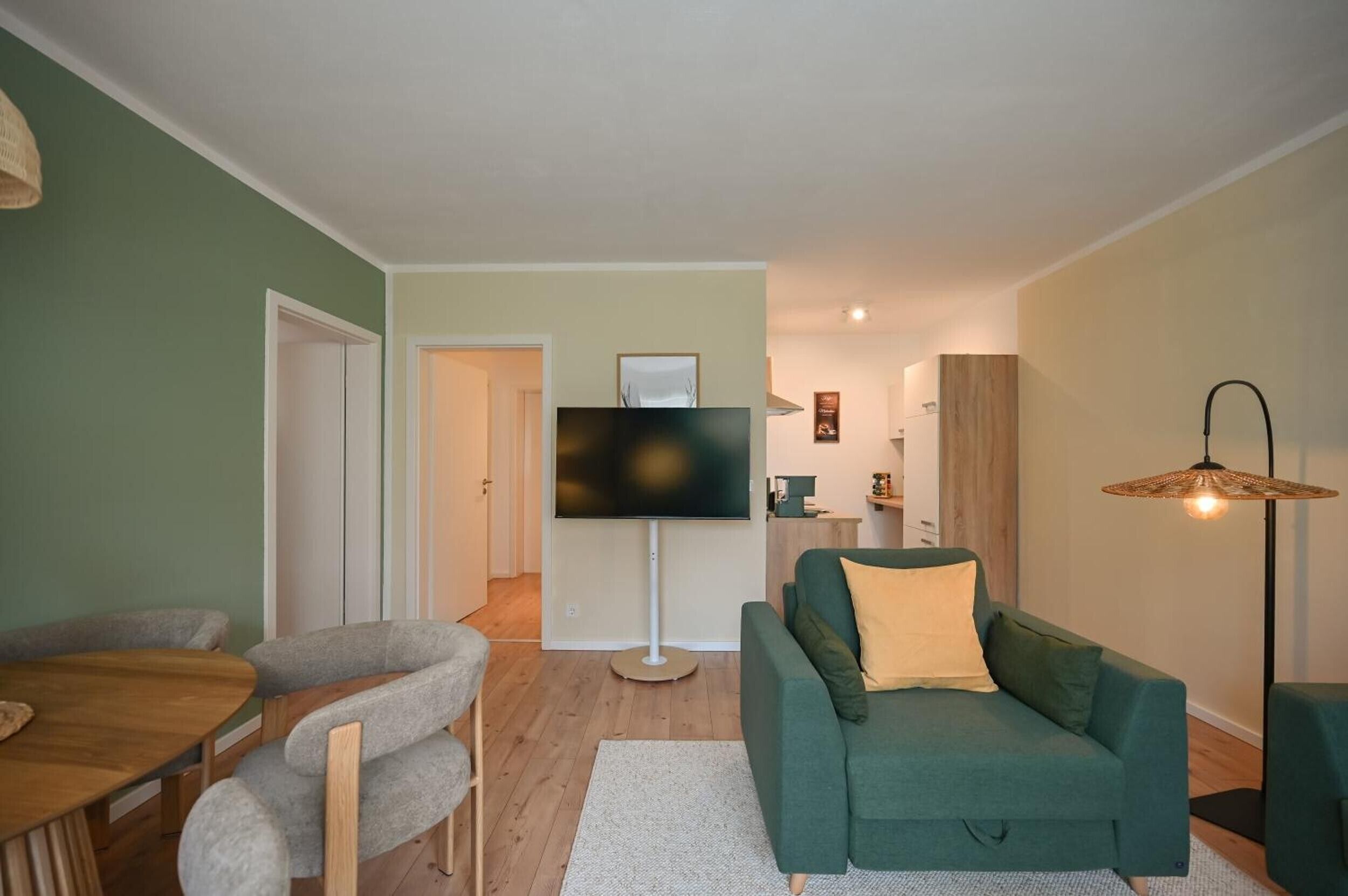 A living room with a green sofa, a flat-screen TV, a dining table with chairs, and a floor lamp.