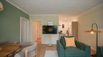A living room with a green sofa, a flat-screen TV, a dining table with chairs, and a floor lamp.