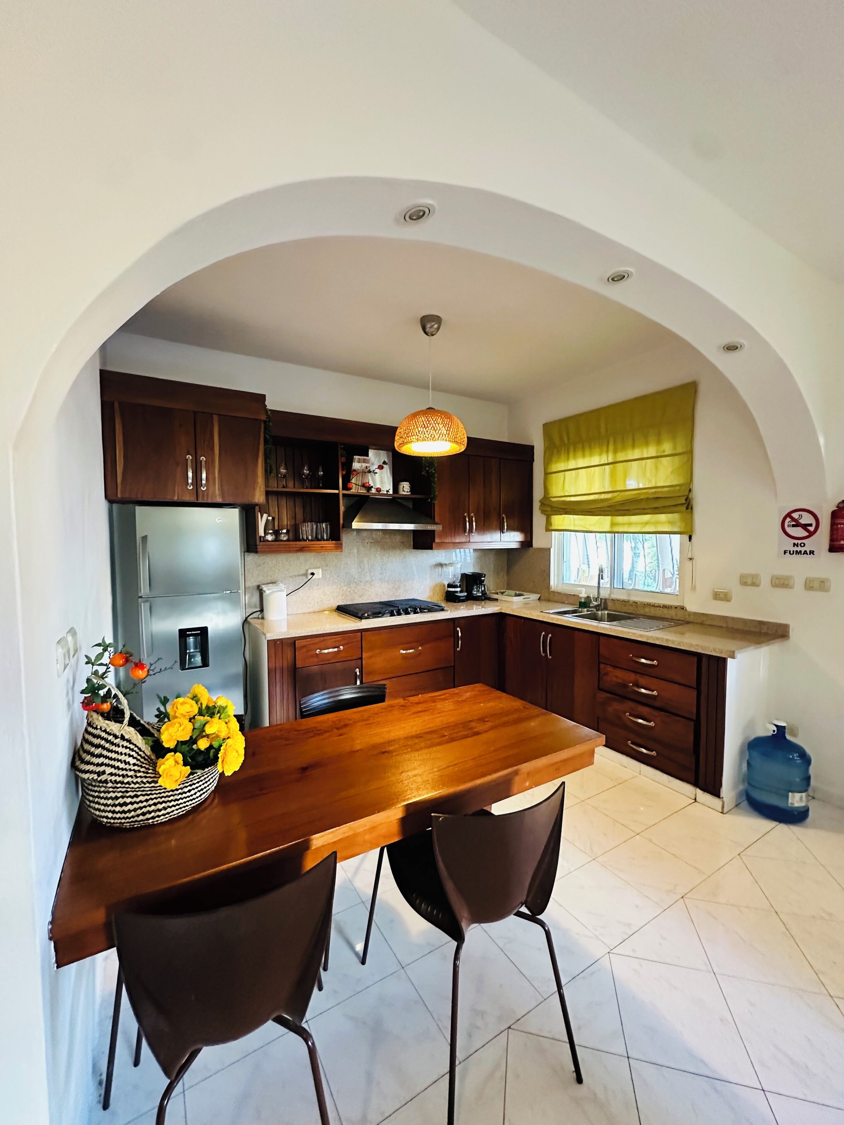 A kitchen with a wooden table, two chairs, a refrigerator, cabinets, and a hanging light fixture.