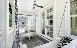Une véranda couverte avec un banc blanc, un ventilateur de plafond et une vue sur l’intérieur de la maison.