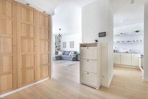 A modern interior with a wooden wardrobe, a grey sofa, and a kitchen area with white cabinets. - Dickensa Apartment (Warszawa)