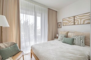 A hotel room with a bed, a chair, a nightstand with a lamp, and a window with curtains. - Dickensa Apartment (Warszawa)