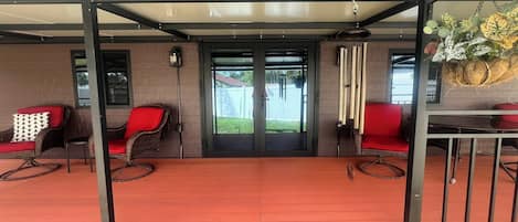 Un espacio de patio techado con sillas de mimbre, una mesa, un muro de vidrio corredizo y una planta colgante.