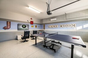 Ein Spielzimmer mit Tischtennisplatte, zwei Gaming-Stühlen, einem Fernseher und einem Projektor.