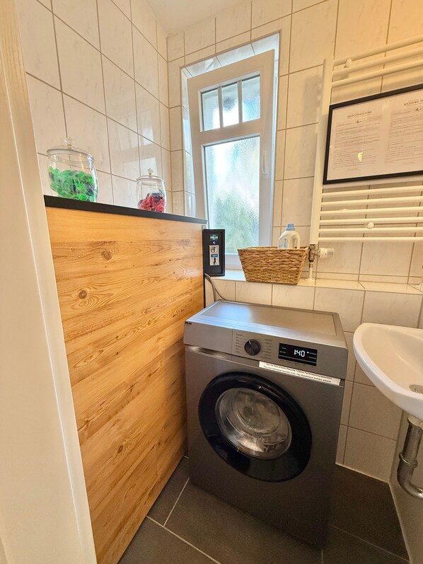 A compact laundry area with a washing machine, a wooden cabinet, a window, and a sink. - Apartment 'Orchidee' with Mini-SPA for 1 Person in Westerwald (Höhr-Grenzhausen)