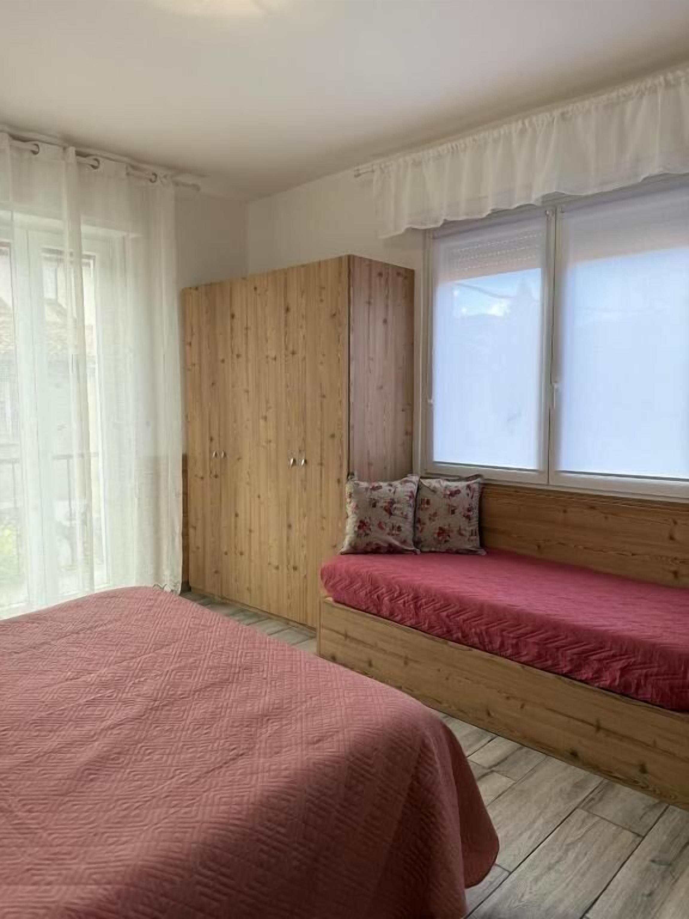 A bedroom with a wooden wardrobe, a bench with a red cushion, and a window with curtains.