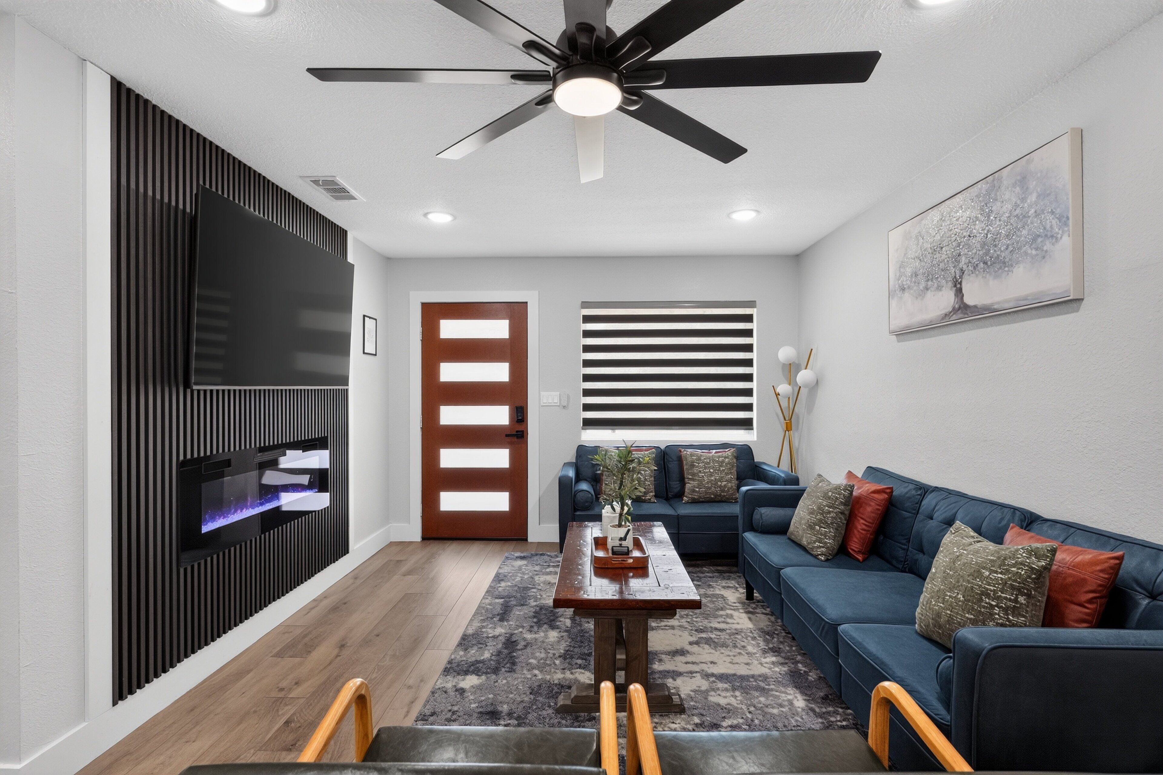 A modern living room with a ceiling fan, a flat-screen TV mounted on a wall, a wooden dining table, a blue sofa with cushions, and a large window with blinds.