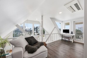 A modern loft bedroom with a sloped ceiling, large windows, a white desk, and a comfortable seating area with a sofa and cushions. - Dolphin Reach Upper | Steps to Beach + Deck (Emerald Isle)
