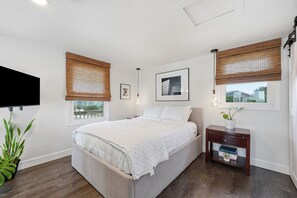 A bedroom with a bed, a bedside table, a TV mounted on the wall, and two windows with bamboo blinds. - Dolphin Reach Upper | Steps to Beach + Deck (Emerald Isle)