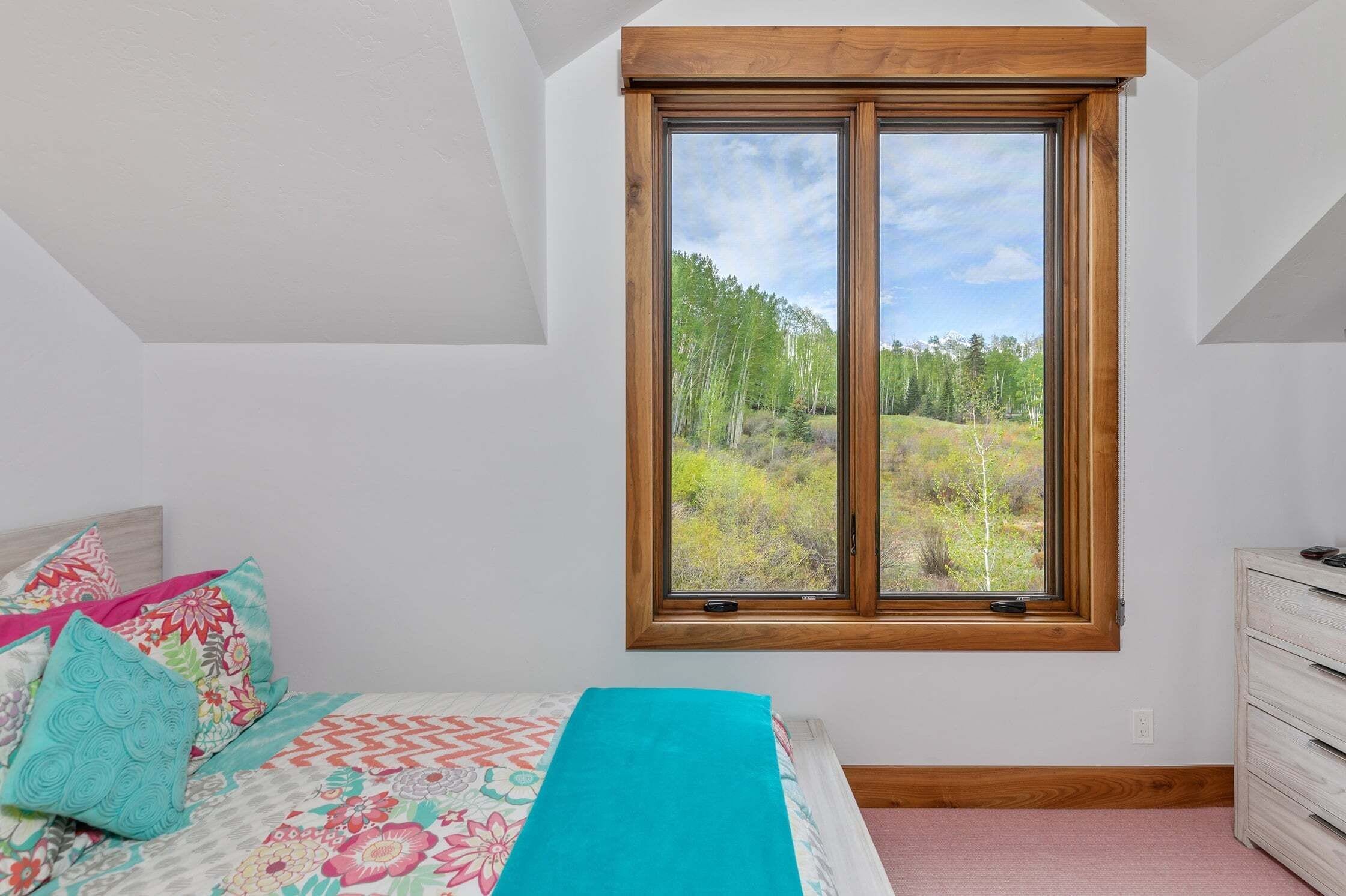 Een slaapkamer met een groot raam, een bed met kleurrijk beddengoed en een dressoir.