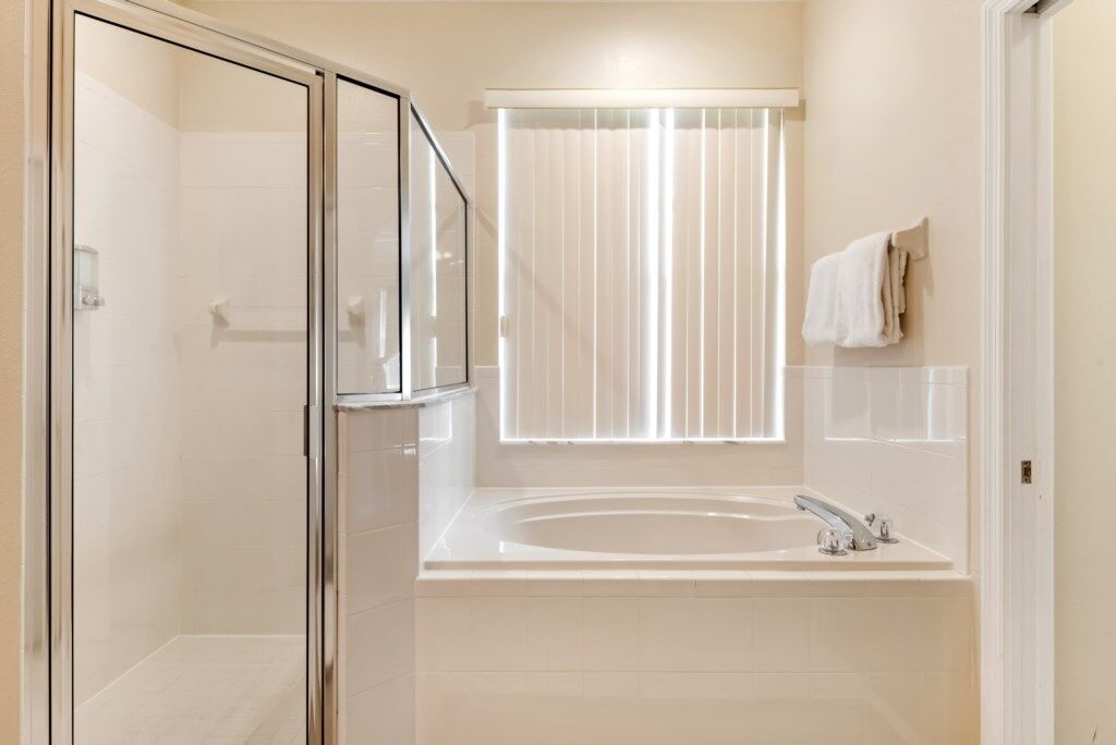 A modern bathroom with a glass shower enclosure, a bathtub, and a window with vertical blinds.