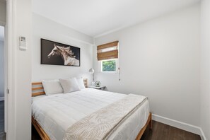 A bedroom with a bed, a nightstand, a lamp, a window with blinds, and a wall-mounted thermostat. - Dolphin Reach Lower | Steps to Beach + Deck (Emerald Isle)