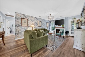 Una sala de estar con un sofá verde, una mesita de centro y un televisor montado en la pared. Hay una chimenea y una ventana con cortinas.