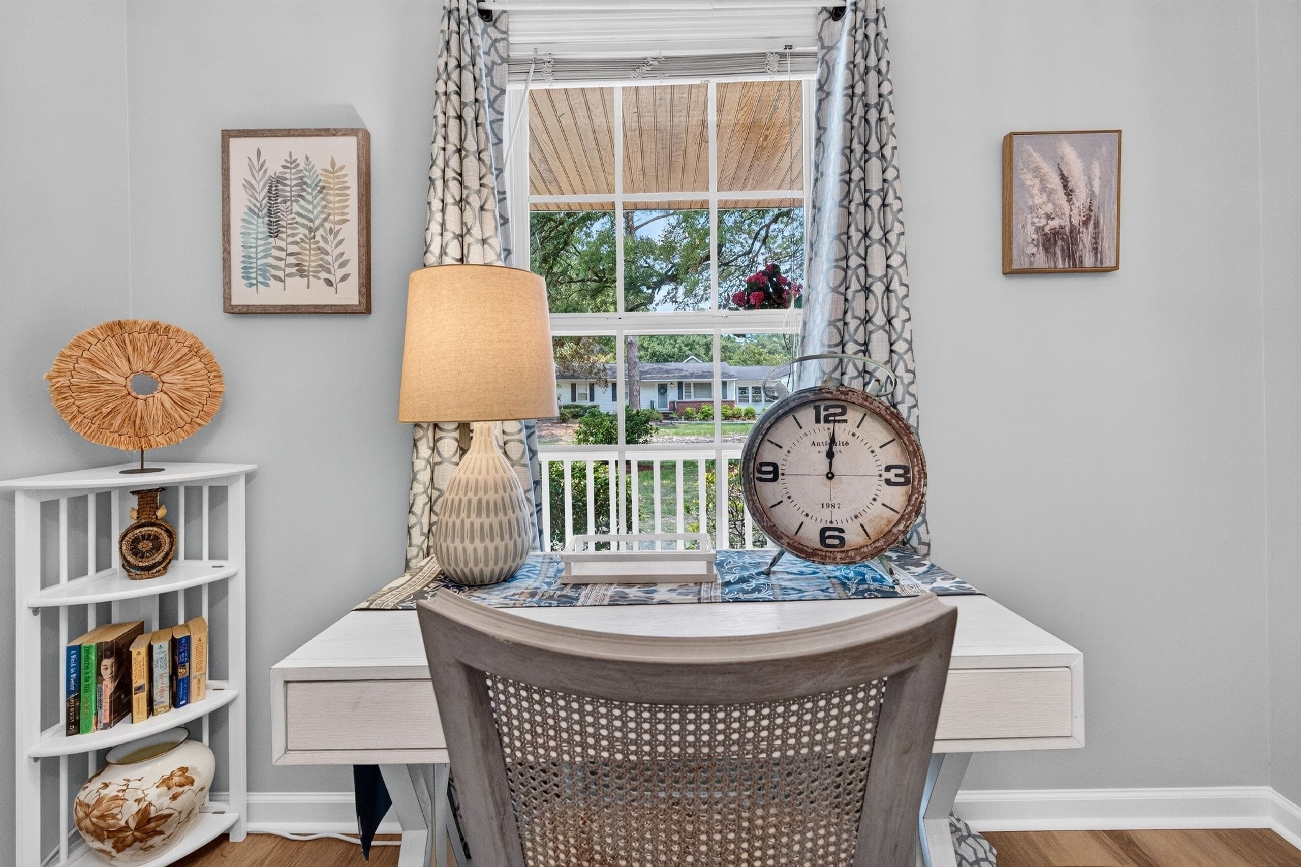 A room with a desk, chair, bookshelves, a lamp, a clock, and framed artwork.
